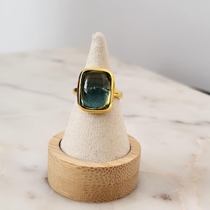 May include: A gold ring with a large, rectangular, green and blue gemstone. The ring is displayed on a small, white, cone-shaped ring holder.