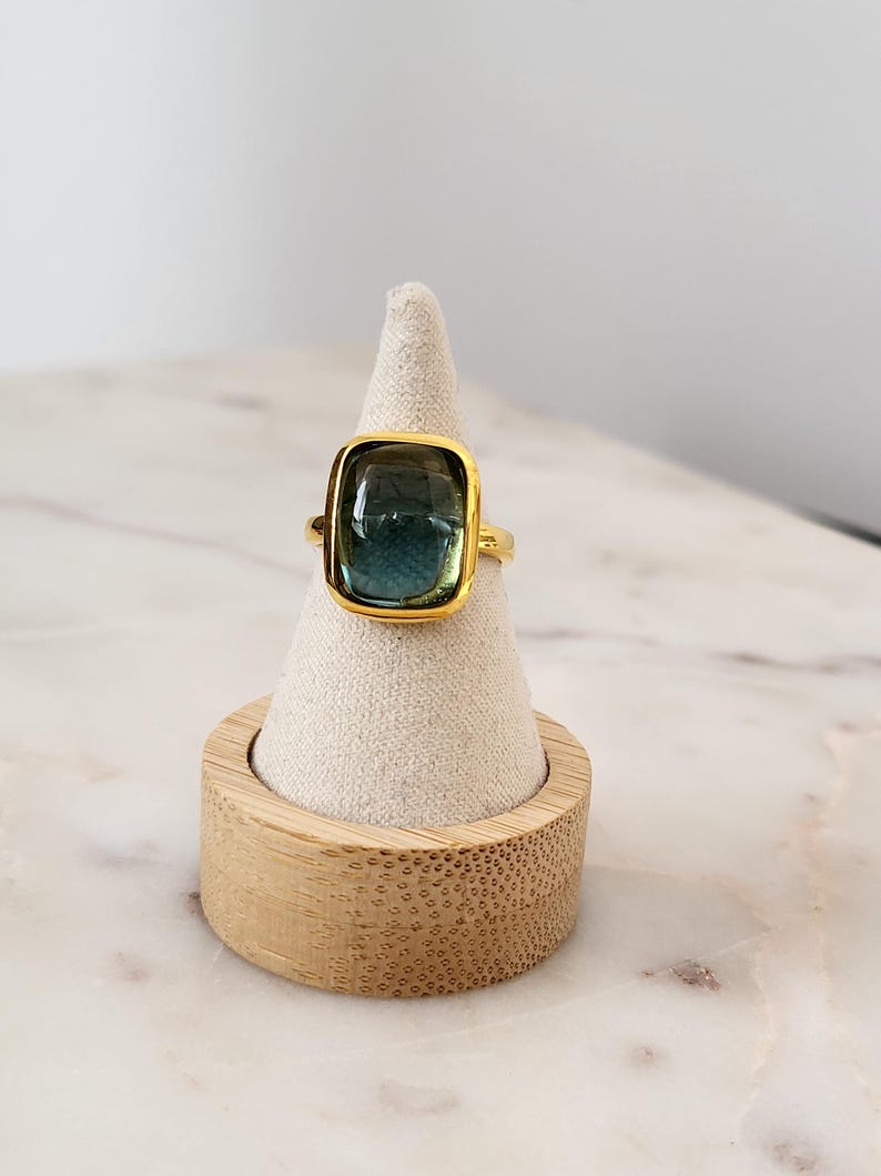 May include: A gold ring with a large, rectangular, green and blue gemstone. The ring is displayed on a small, white, cone-shaped ring holder.