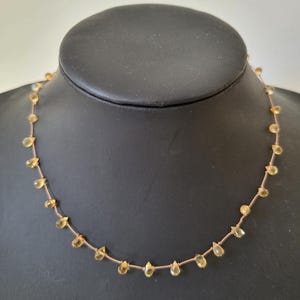 Puede incluir: Un delicado collar con pequeñas piedras preciosas amarillas en forma de lágrima ensartadas en un fino cordón marrón claro. El collar se exhibe en un maniquí negro, mostrando el diseño y el color de la joya.