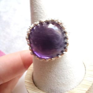 May include: A large, round amethyst ring with a silver-colored, ornate setting. The deep purple gemstone is the focal point, set against a light background. The ring is displayed on a ring holder.