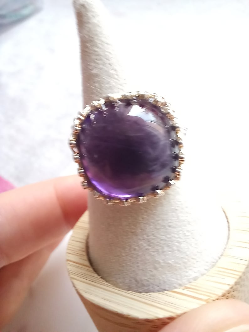 May include: A large, round amethyst ring with a silver-colored, ornate setting. The deep purple gemstone is the focal point, set against a light background. The ring is displayed on a ring holder.
