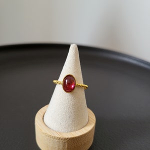Puede incluir: Un anillo de oro con una piedra preciosa roja engastada en un engaste de bisel. El anillo est&aacute; en un expositor de anillo de tela blanca.