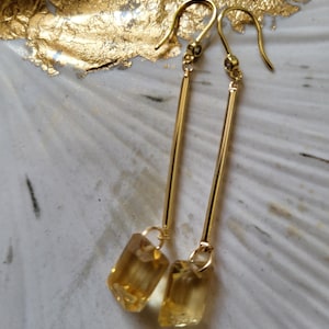 May include: A pair of gold-tone drop earrings featuring a long, slender metal bar suspended from a hook wire. At the base of each bar hangs a rectangular, faceted yellow gemstone that catches the light, creating a clear and polished appearance.