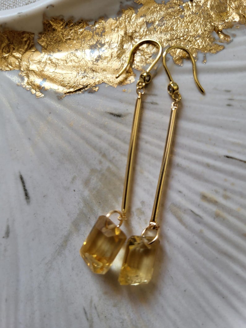 May include: A pair of gold-tone drop earrings featuring a long, slender metal bar suspended from a hook wire. At the base of each bar hangs a rectangular, faceted yellow gemstone that catches the light, creating a clear and polished appearance.