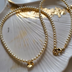 Peut inclure: Un collier de perles délicat avec un fermoir doré et un seul pendentif en forme de goutte, de couleur ambre. Le collier est présenté sur un plat blanc en forme de coquillage avec des accents dorés.