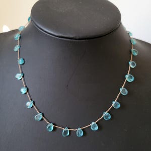 Pu&ograve; includere: Una delicata collana con pietre preziose a forma di goccia, di colore azzurro chiaro, infilate su una sottile catena color oro. La collana &egrave; esposta su un manichino nero, che mette in risalto il design e il colore del gioiello.