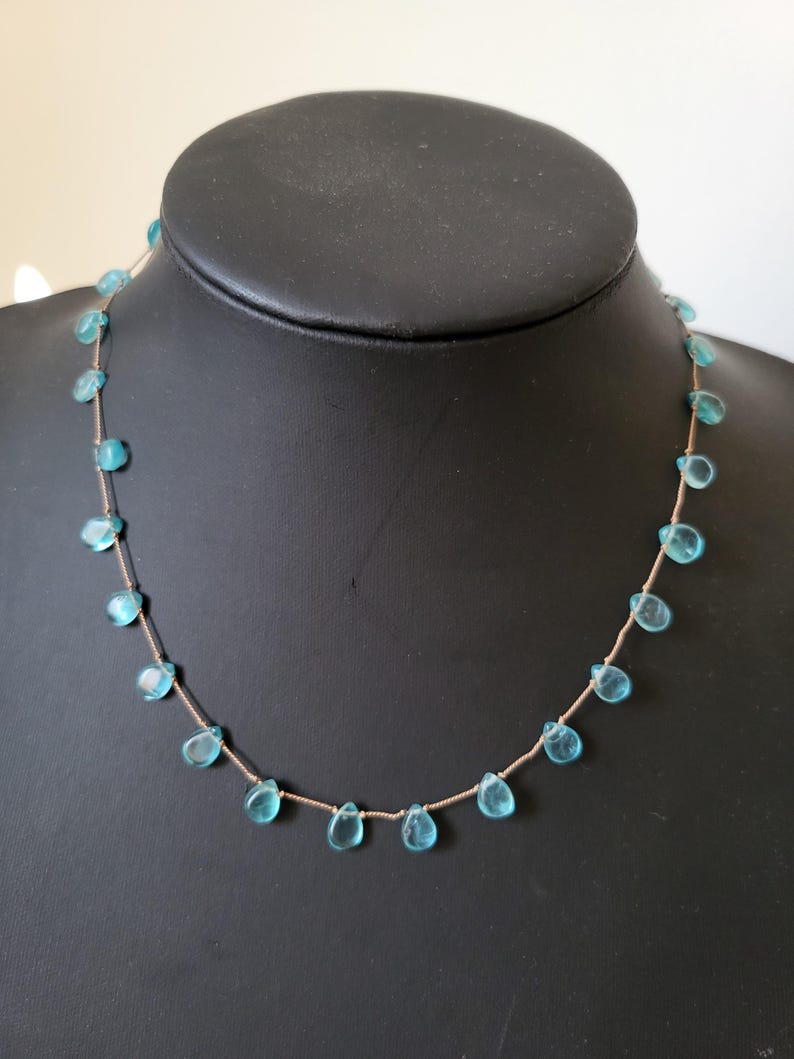 Pu&ograve; includere: Una delicata collana con pietre preziose a forma di goccia, di colore azzurro chiaro, infilate su una sottile catena color oro. La collana &egrave; esposta su un manichino nero, che mette in risalto il design e il colore del gioiello.