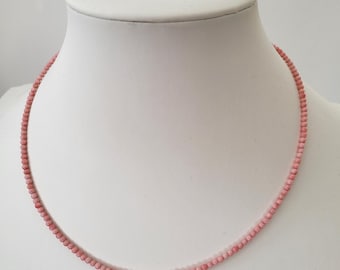 Puede incluir: Un delicado collar con pequeñas cuentas redondas de color rosa claro. Las cuentas están ensartadas para formar un collar de una sola hebra. El collar se exhibe en un busto de maniquí blanco, mostrando su simple elegancia.