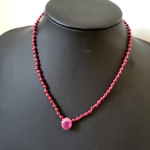 May include: A ruby necklace displayed on a black mannequin. The necklace features small, round ruby beads strung together, with a larger, oval-shaped ruby pendant at the center. The beads and pendant are a deep, rich red color.