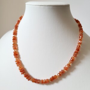 Peut inclure: Un collier compos&eacute; de perles de soleil orange-rouge facett&eacute;es. Les perles sont enfil&eacute;es ensemble, cr&eacute;ant un bijou d&eacute;licat et scintillant. Le collier est pr&eacute;sent&eacute; sur un buste de mannequin blanc, mettant en valeur son design et sa couleur.