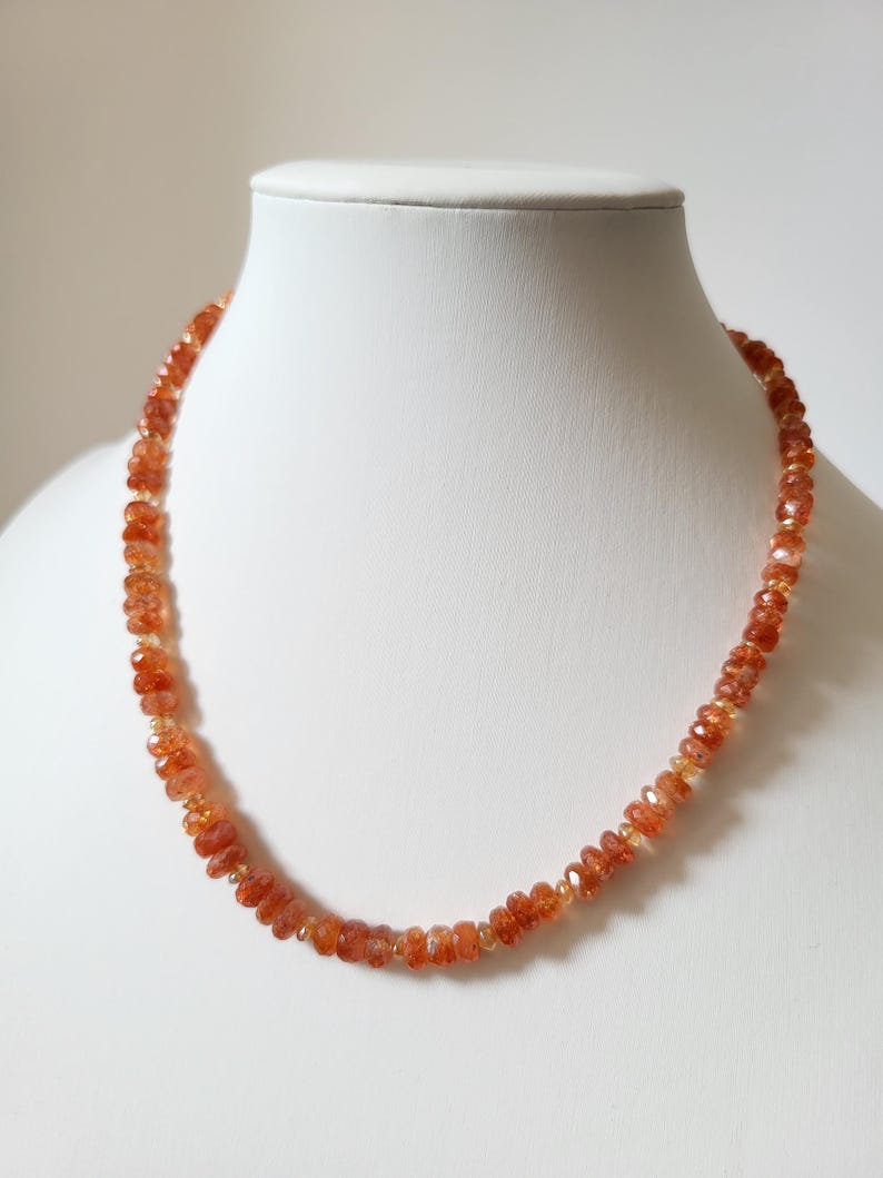 Peut inclure: Un collier compos&eacute; de perles de soleil orange-rouge facett&eacute;es. Les perles sont enfil&eacute;es ensemble, cr&eacute;ant un bijou d&eacute;licat et scintillant. Le collier est pr&eacute;sent&eacute; sur un buste de mannequin blanc, mettant en valeur son design et sa couleur.
