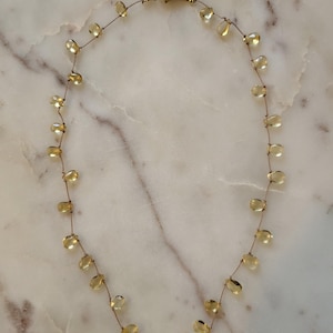 May include: A delicate necklace featuring a thin, brown cord strung with numerous small, translucent, pale yellow, teardrop-shaped beads spaced evenly along the length. The necklace is secured with a small, gold-toned metal lobster claw clasp.
