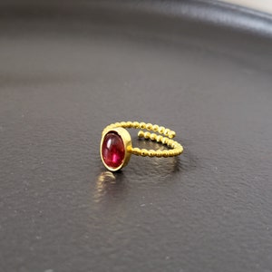 Puede incluir: Un anillo de oro con una piedra ovalada roja engastada en el centro. La banda del anillo est&aacute; hecha de peque&ntilde;as cuentas doradas.