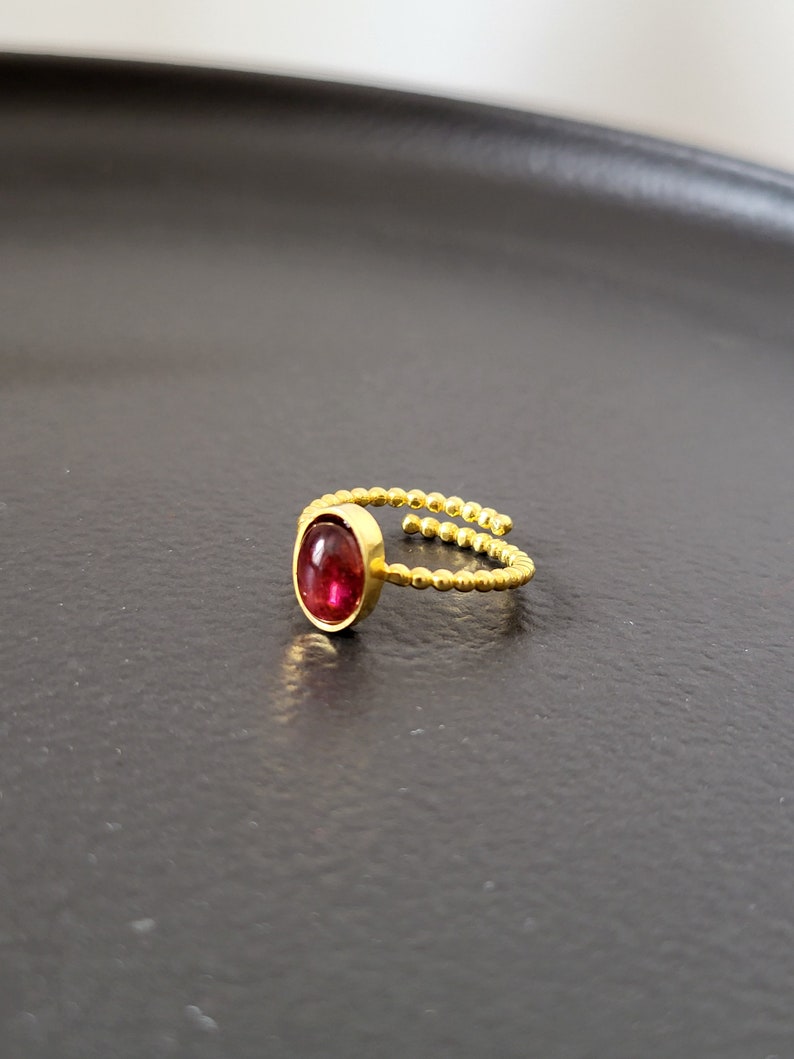 Puede incluir: Un anillo de oro con una piedra ovalada roja engastada en el centro. La banda del anillo est&aacute; hecha de peque&ntilde;as cuentas doradas.