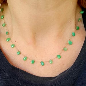 May include: A delicate necklace featuring small, teardrop-shaped green gemstones strung on a thin, gold-colored chain. The necklace is displayed against a dark background, highlighting the vibrant green color of the stones.