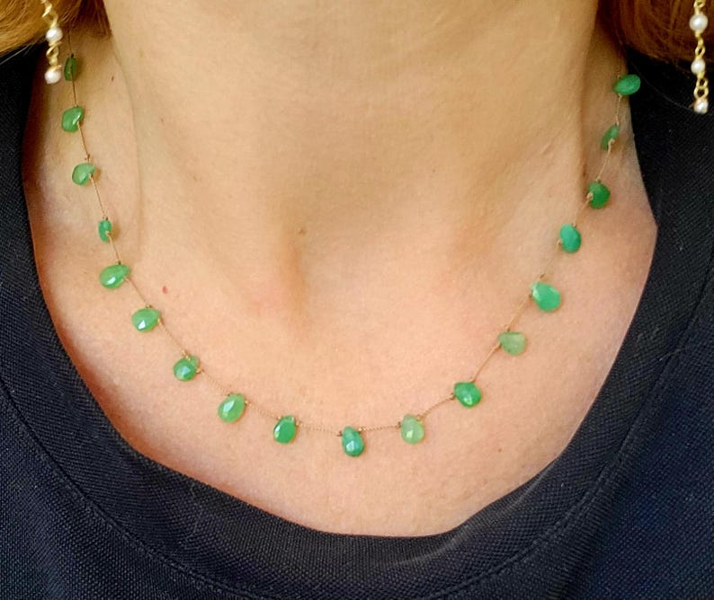 May include: A delicate necklace featuring small, teardrop-shaped green gemstones strung on a thin, gold-colored chain. The necklace is displayed against a dark background, highlighting the vibrant green color of the stones.