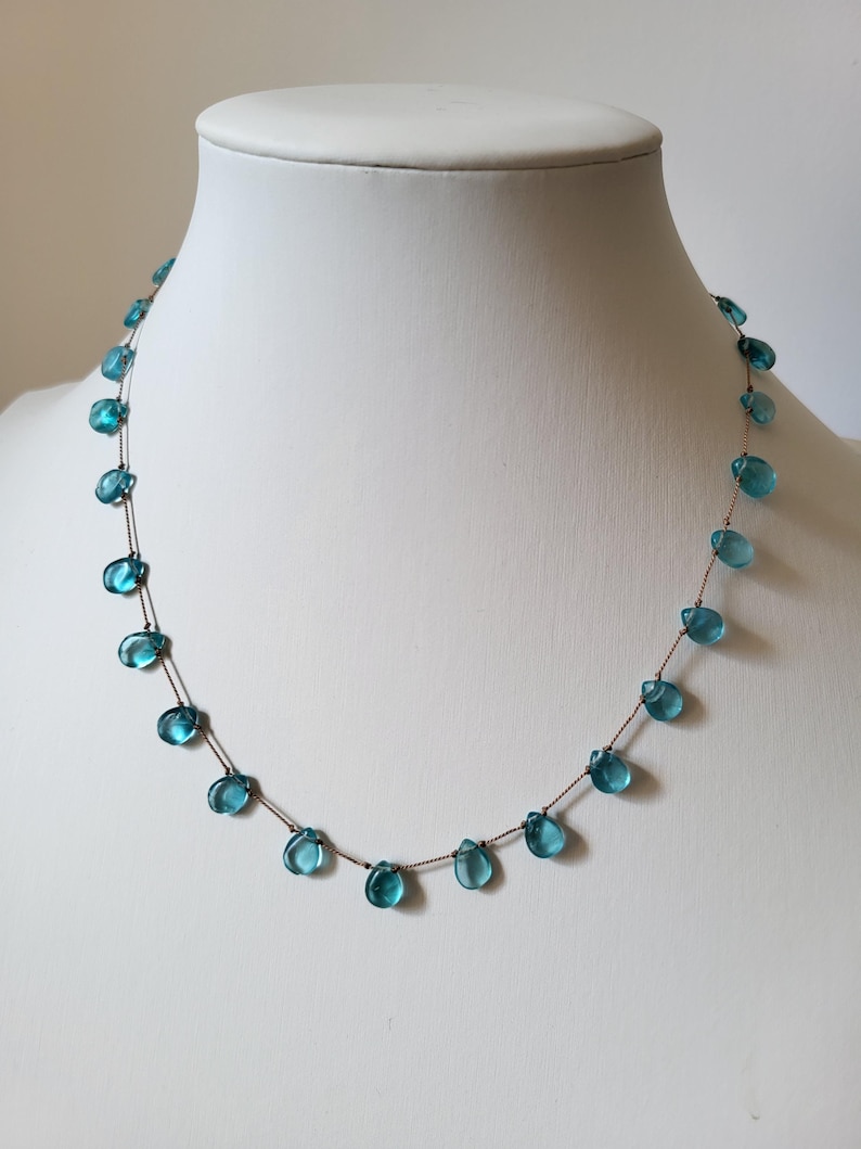 Pu&ograve; includere: Una delicata collana con perline a forma di goccia, di colore azzurro chiaro, infilate su una sottile catena marrone. Le perline sono distanziate in modo uniforme, creando un design minimalista ed elegante. La collana &egrave; esposta su un manichino bianco.