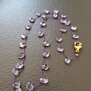 May include: A delicate necklace featuring clear, faceted teardrop-shaped beads strung on a vibrant purple cord. The necklace is secured with a gold-colored clasp. The beads are evenly spaced along the cord, creating a simple yet elegant design.