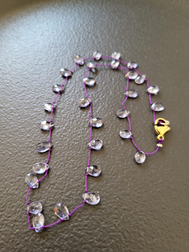 May include: A delicate necklace featuring clear, faceted teardrop-shaped beads strung on a vibrant purple cord. The necklace is secured with a gold-colored clasp. The beads are evenly spaced along the cord, creating a simple yet elegant design.