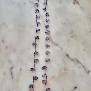 May include: Delicate necklace featuring teardrop-shaped, translucent blue-gray beads strung on a dark purple cord. The necklace has a gold-colored clasp. The beads are evenly spaced along the cord, creating a simple yet elegant design.