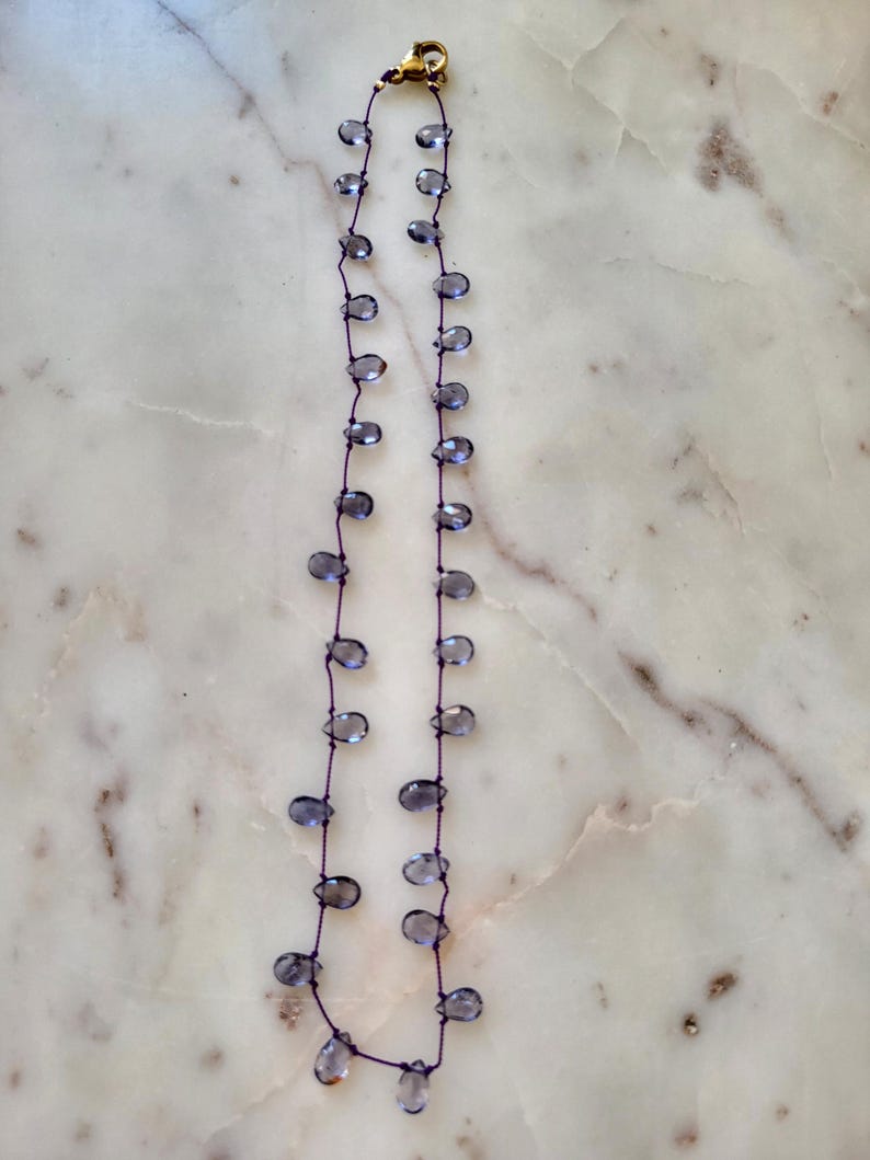 May include: Delicate necklace featuring teardrop-shaped, translucent blue-gray beads strung on a dark purple cord. The necklace has a gold-colored clasp. The beads are evenly spaced along the cord, creating a simple yet elegant design.
