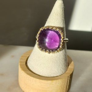 May include: A square-cut, purple gemstone ring with a gold and silver band. The ring is displayed on a white, cone-shaped stand, set on a wooden base. The gemstone is a deep, vibrant purple.