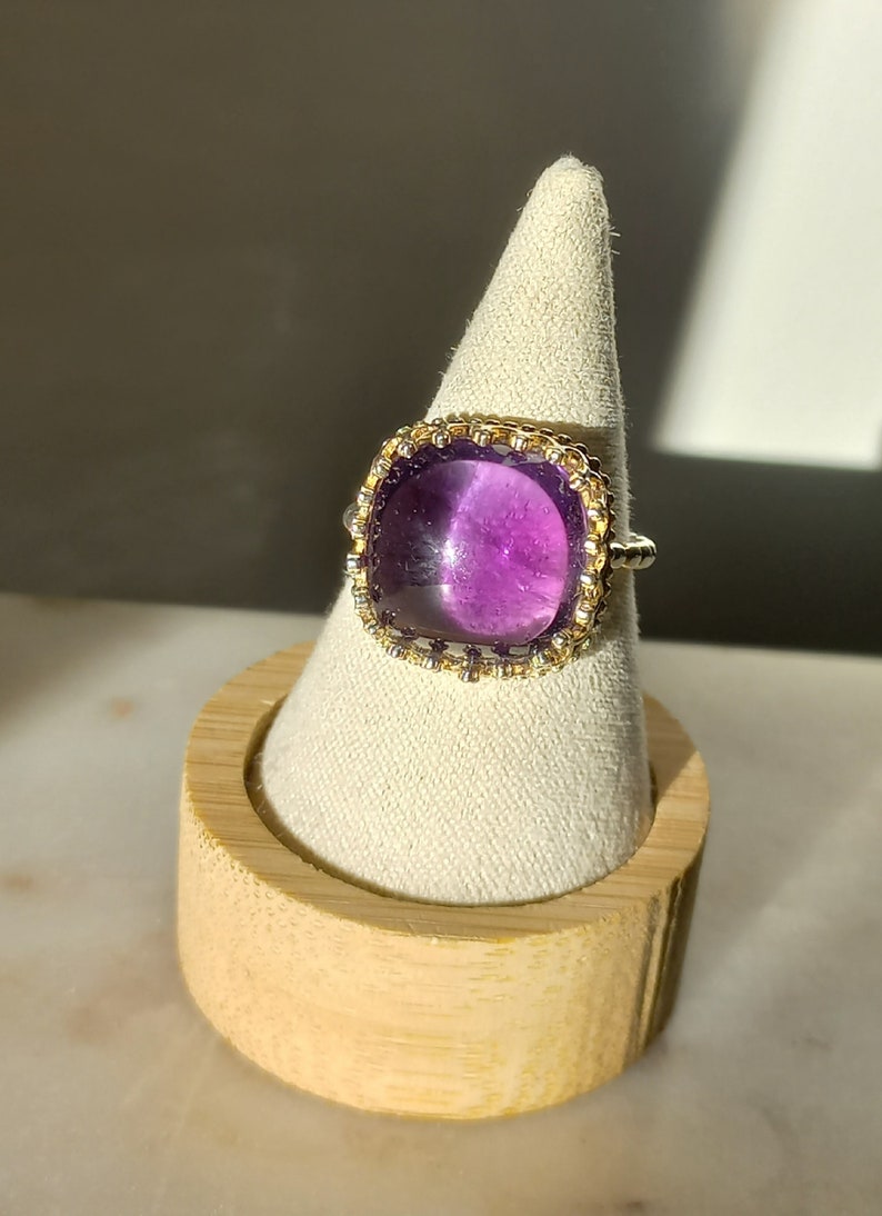 May include: A square-cut, purple gemstone ring with a gold and silver band. The ring is displayed on a white, cone-shaped stand, set on a wooden base. The gemstone is a deep, vibrant purple.