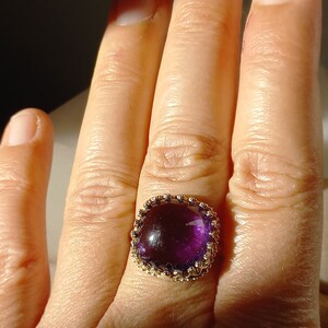 May include: A gold ring featuring a large, square-cut, deep purple gemstone. The gemstone is set in a decorative gold band with a detailed, ornate design. The ring is shown on a person's finger.