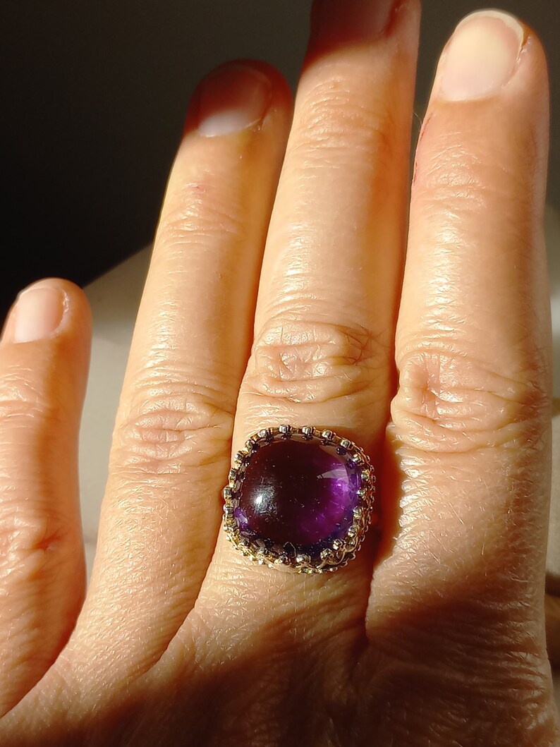 May include: A gold ring featuring a large, square-cut, deep purple gemstone. The gemstone is set in a decorative gold band with a detailed, ornate design. The ring is shown on a person's finger.
