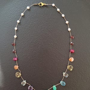 Op de afbeelding: Een delicate ketting met een dun koord geregen met kleine witte parels en een verscheidenheid aan veelkleurige, druppelvormige gefacetteerde edelstenen. De ketting wordt vastgezet met een goudkleurige metalen karabijnsluiting.