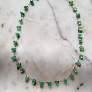 Pode incluir: Um delicado colar de pedras preciosas verdes com um design simples. O colar apresenta pequenas pedras verdes em forma de l&aacute;grima enfiadas em um cord&atilde;o marrom fino.