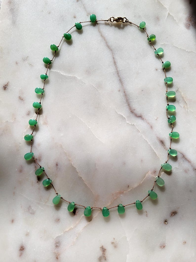 Pode incluir: Um delicado colar de pedras preciosas verdes com um design simples. O colar apresenta pequenas pedras verdes em forma de l&aacute;grima enfiadas em um cord&atilde;o marrom fino.