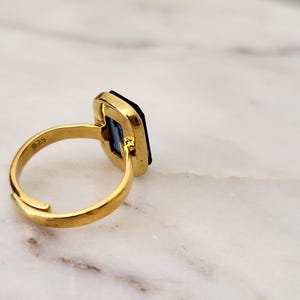 May include: A gold-colored ring with a rectangular, dark blue gemstone. The ring has a simple band and a setting that frames the stone. The ring is set against a white and gray marble surface. The ring is marked with "925".