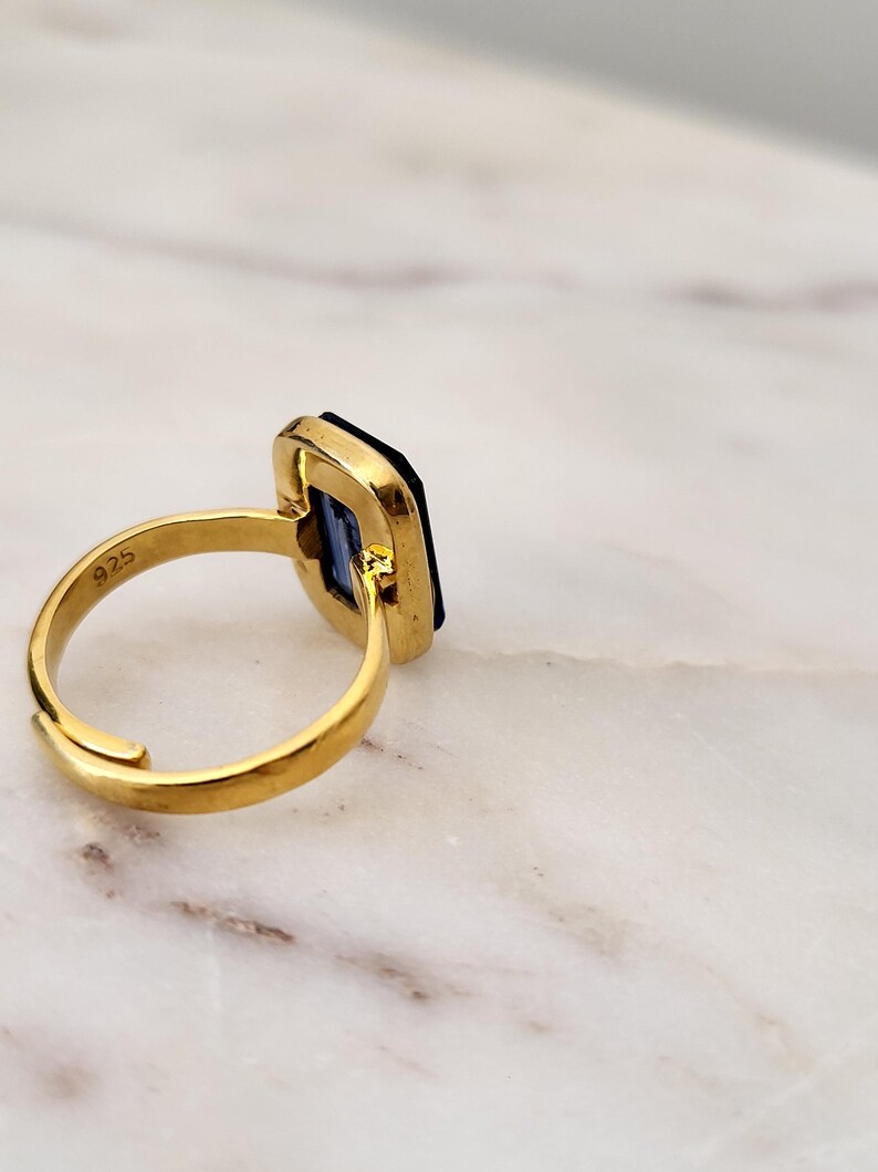 May include: A gold-colored ring with a rectangular, dark blue gemstone. The ring has a simple band and a setting that frames the stone. The ring is set against a white and gray marble surface. The ring is marked with "925".