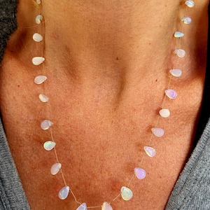 May include: A delicate necklace featuring teardrop-shaped opals strung on a thin, gold-colored chain. The opals display a range of soft colors, including white, pink, and hints of blue, creating a subtle, iridescent effect. The necklace is designed to be worn close to the neck.