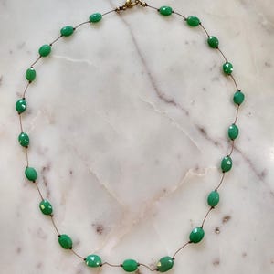 May include: A delicate necklace featuring oval-shaped green beads strung on a thin, brown cord. The beads have a slightly faceted appearance, and the necklace is secured with a gold-colored clasp. The necklace is displayed on a white and gray marble surface.