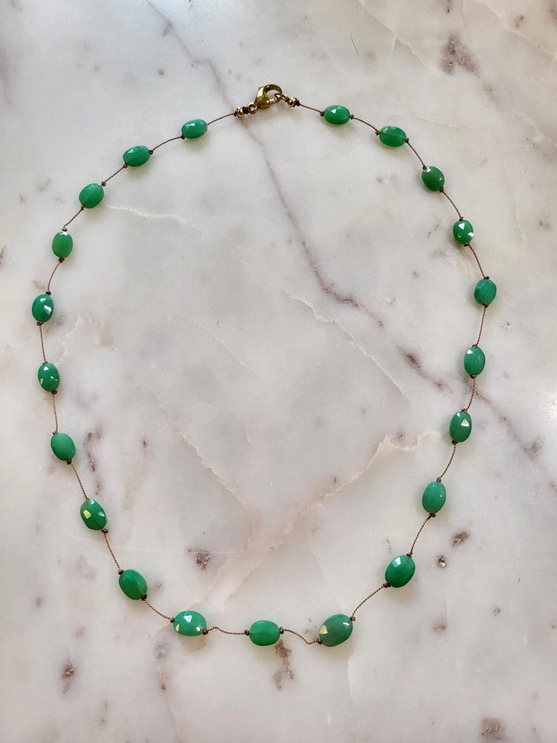 May include: A delicate necklace featuring oval-shaped green beads strung on a thin, brown cord. The beads have a slightly faceted appearance, and the necklace is secured with a gold-colored clasp. The necklace is displayed on a white and gray marble surface.