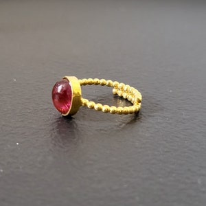 May include: A gold ring with a pink gemstone set in a bezel setting. The band is made of small gold beads.