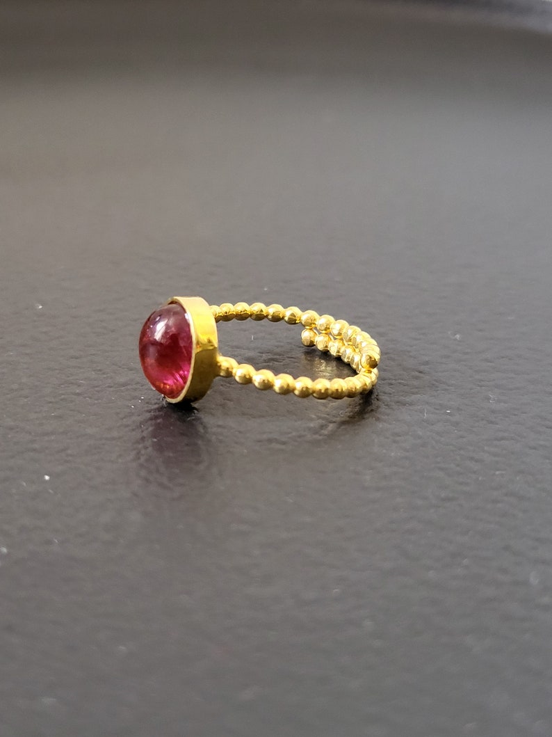 May include: A gold ring with a pink gemstone set in a bezel setting. The band is made of small gold beads.