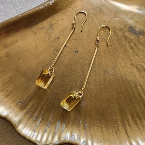 May include: A pair of gold-tone drop earrings featuring long, slender metal bars suspended from curved ear wires. Each earring terminates in a rectangular, faceted yellow gemstone that catches the light, creating a clear and elegant design.