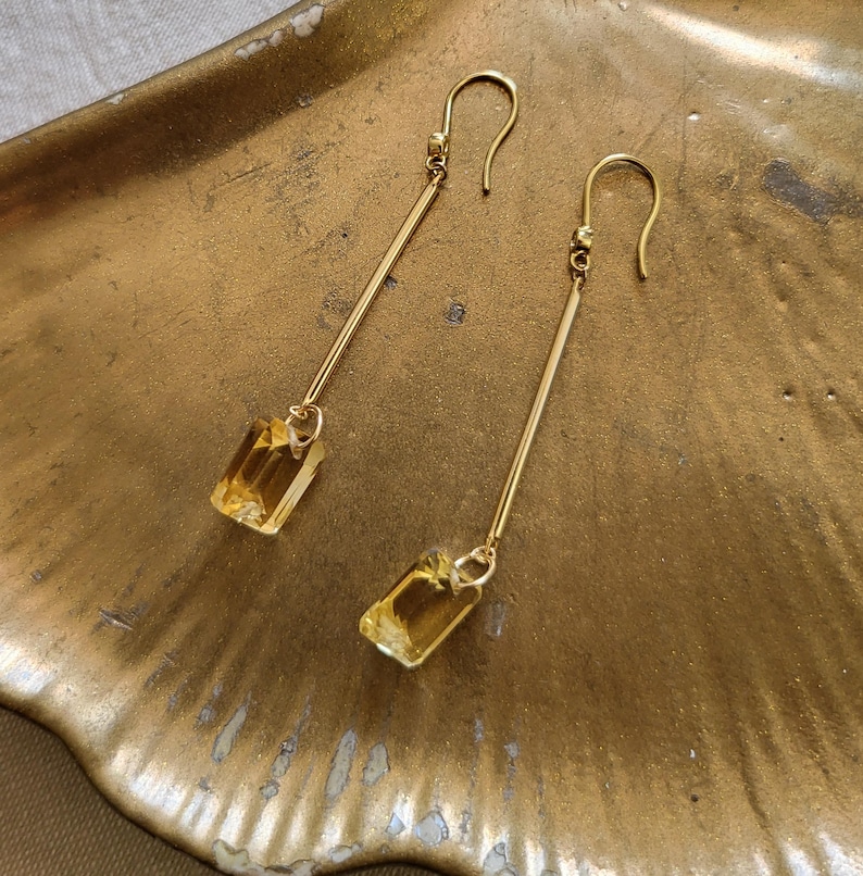 May include: A pair of gold-tone drop earrings featuring long, slender metal bars suspended from curved ear wires. Each earring terminates in a rectangular, faceted yellow gemstone that catches the light, creating a clear and elegant design.