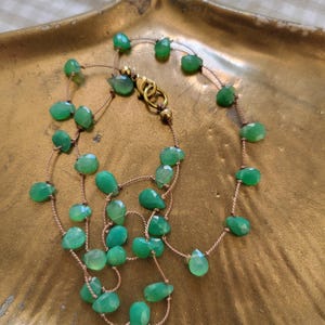 May include: A delicate necklace featuring faceted, light green gemstones strung on a thin, tan cord. The necklace is secured with a gold-colored clasp. The gemstones are irregularly shaped and vary in size, creating a unique, organic look. The necklace rests on a gold-toned, leaf-shaped dish.