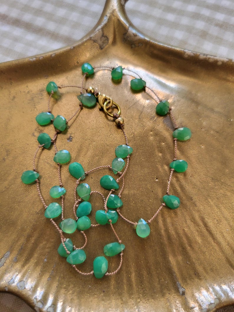 May include: A delicate necklace featuring faceted, light green gemstones strung on a thin, tan cord. The necklace is secured with a gold-colored clasp. The gemstones are irregularly shaped and vary in size, creating a unique, organic look. The necklace rests on a gold-toned, leaf-shaped dish.