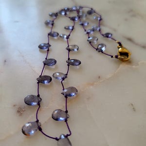 May include: A delicate necklace featuring teardrop-shaped, translucent blue-gray beads strung on a thin, dark purple cord. The necklace has a gold-colored clasp. The beads are evenly spaced, creating a graceful, flowing design.