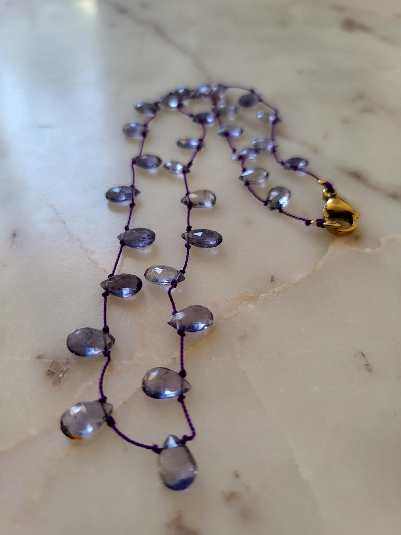 May include: A delicate necklace featuring teardrop-shaped, translucent blue-gray beads strung on a thin, dark purple cord. The necklace has a gold-colored clasp. The beads are evenly spaced, creating a graceful, flowing design.