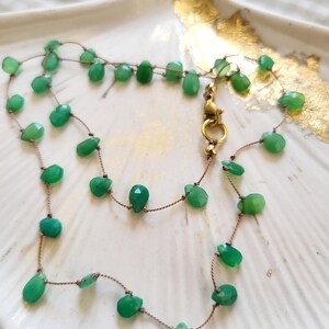 May include: A delicate necklace featuring faceted green gemstone beads strung on a thin, brown cord. The necklace has a gold-colored clasp and is displayed on a white, scalloped dish with gold accents. The gemstones are various shapes and sizes.