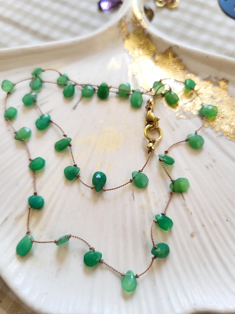 May include: A delicate necklace featuring faceted green gemstone beads strung on a thin, brown cord. The necklace has a gold-colored clasp and is displayed on a white, scalloped dish with gold accents. The gemstones are various shapes and sizes.