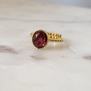 May include: A gold ring with a pink oval gemstone set in the center. The ring band is made of small gold beads.
