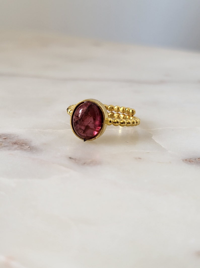May include: A gold ring with a pink oval gemstone set in the center. The ring band is made of small gold beads.