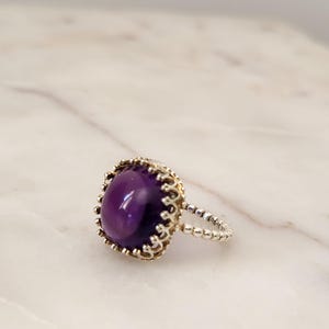 May include: A silver ring with a large, oval, deep purple gemstone. The gemstone is set in a gold-colored, ornate bezel. The ring band is silver and features a beaded design. The ring is displayed on a white marble surface.